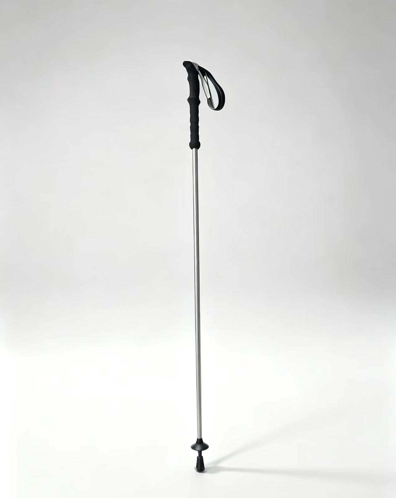 A single hiking pole standing upright on a clean light gray studio background. The pole has a silver aluminum shaft with black accents on the top loop, handle, and tip.