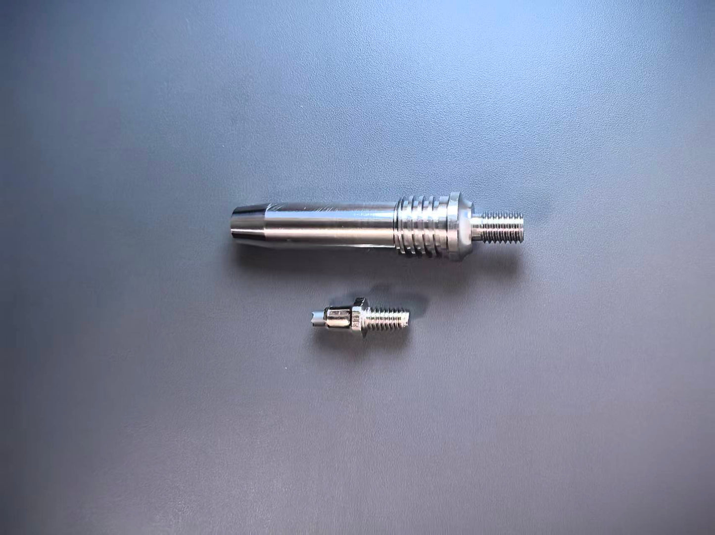 A pair of machined metal threaded components, including a larger cylindrical sleeve with a threaded end and a smaller matching threaded piece, displayed on a smooth gray surface.