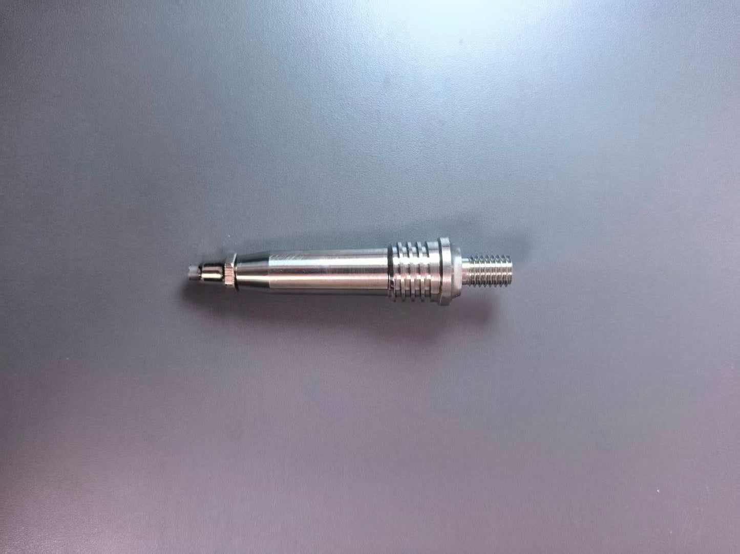 A metallic cylindrical mechanical part with a threaded end and a textured ridged section, photographed on a plain gray surface.