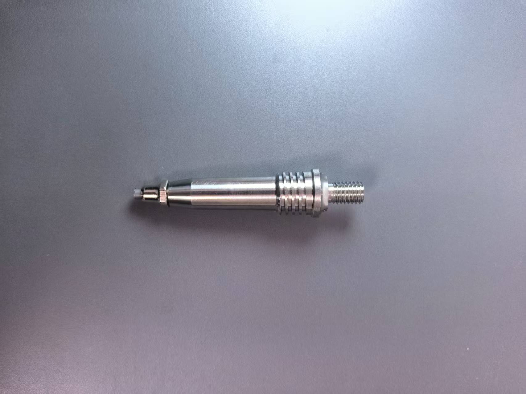 A metallic cylindrical mechanical part with a threaded end and a textured ridged section, photographed on a plain gray surface.