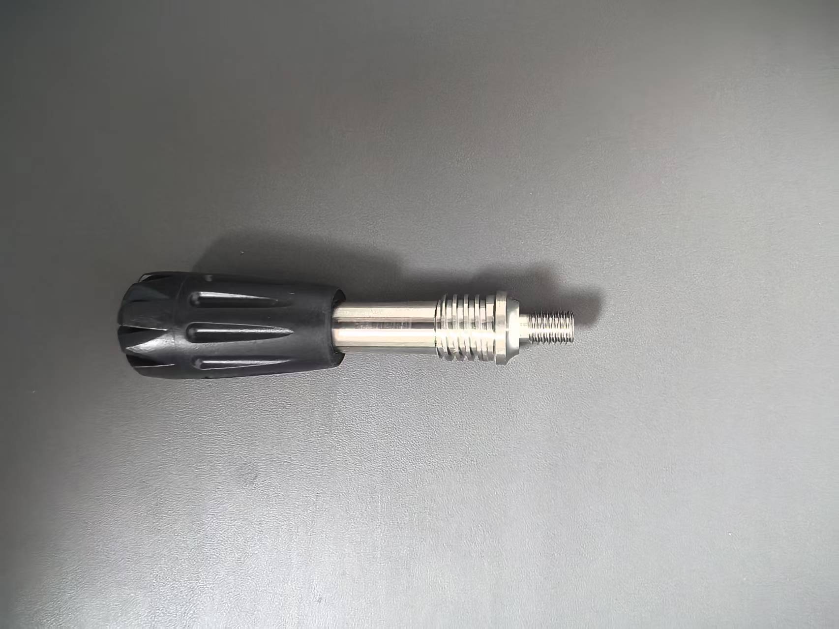 A replacement knob or handle with a black ribbed plastic head and a silver metal threaded shaft, shown on a plain gray surface.