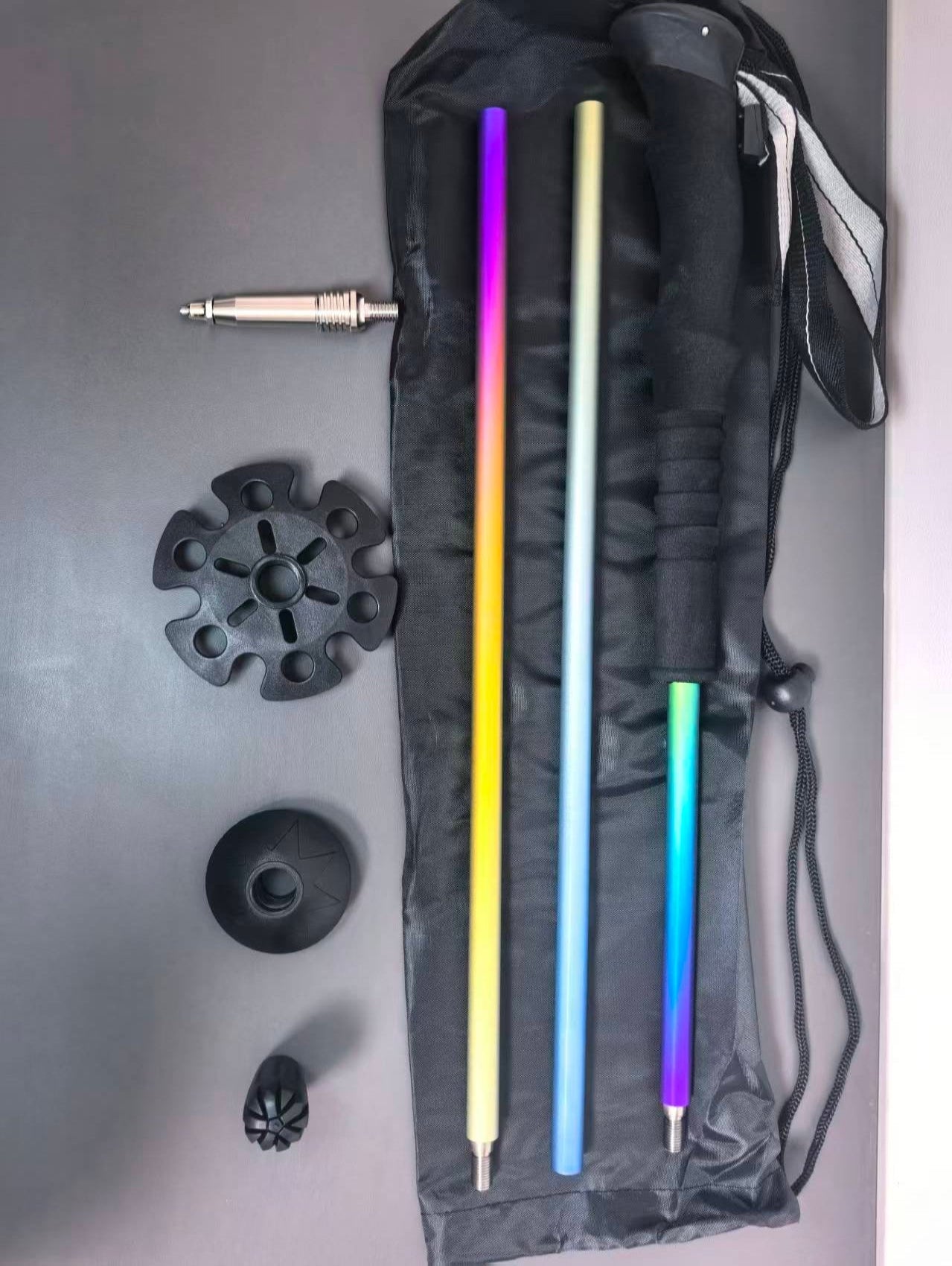 A set of three telescoping walking poles with vibrant rainbow gradient handles, laid out next to their black carrying case on a gray surface. The set includes a black multi-directional rubberized foot plate, a black wrist strap, and a silver metal quick-release connector.
